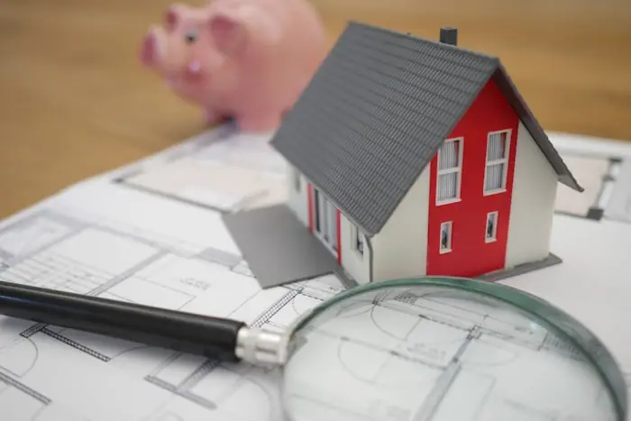 Model house on building plans beside a magnifying glass and a coin bank